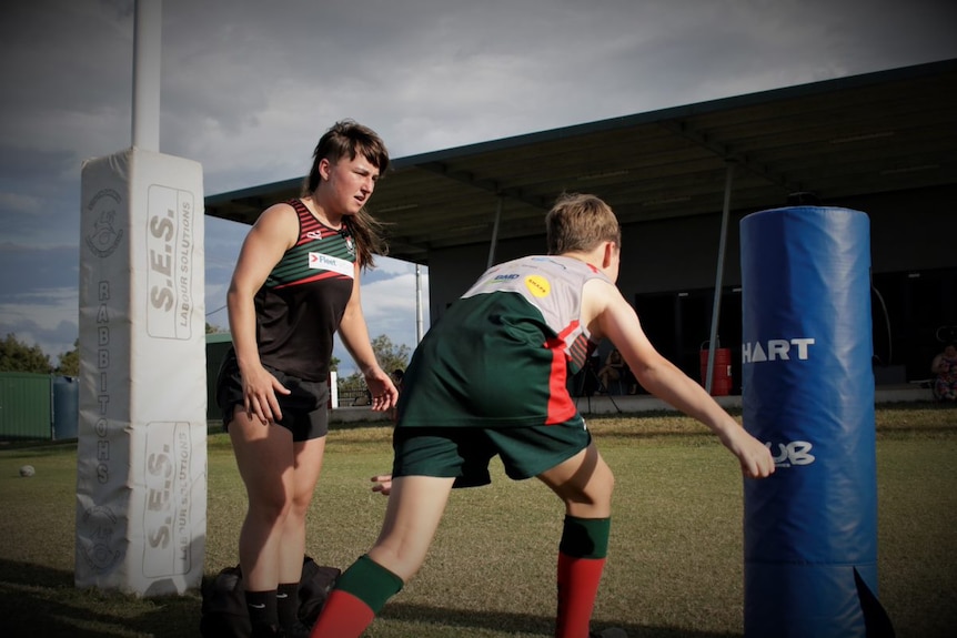 A young woman concentrates on training a young boy rugby skills at an oval. 