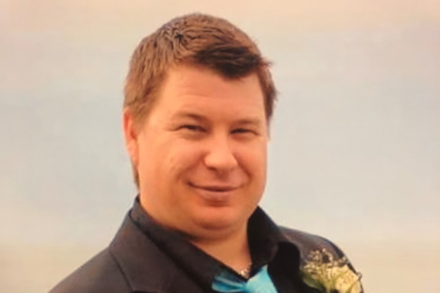 A man smiles at the camera in a black suit with an aqua blue tie