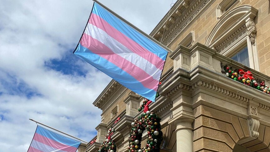 Pale blue and pale pink striped flags fly outside a heritage sandstone building 