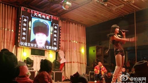 A stripper is on stage in front of a screen with a photo of the deceased.