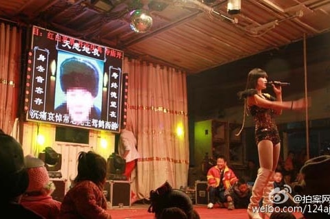 A stripper is on stage in front of a screen with a photo of the deceased.