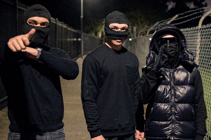 Three young men in black stand wearing masks and balaclavas. They are making signals with their hands.