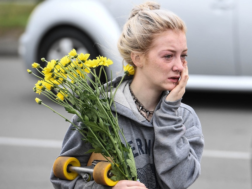 A woman carrying yellow flowers and a skateboard wipes away tears as she walks to join a cleansing ceremony outside a mosque