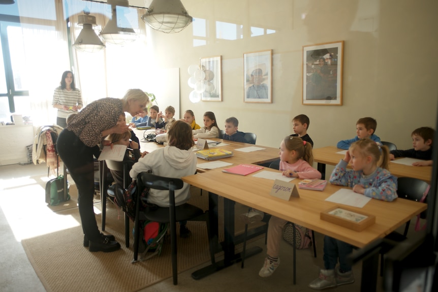 a teacher leans over a desk speaking to students in a classroom with a big window letting in lots of light