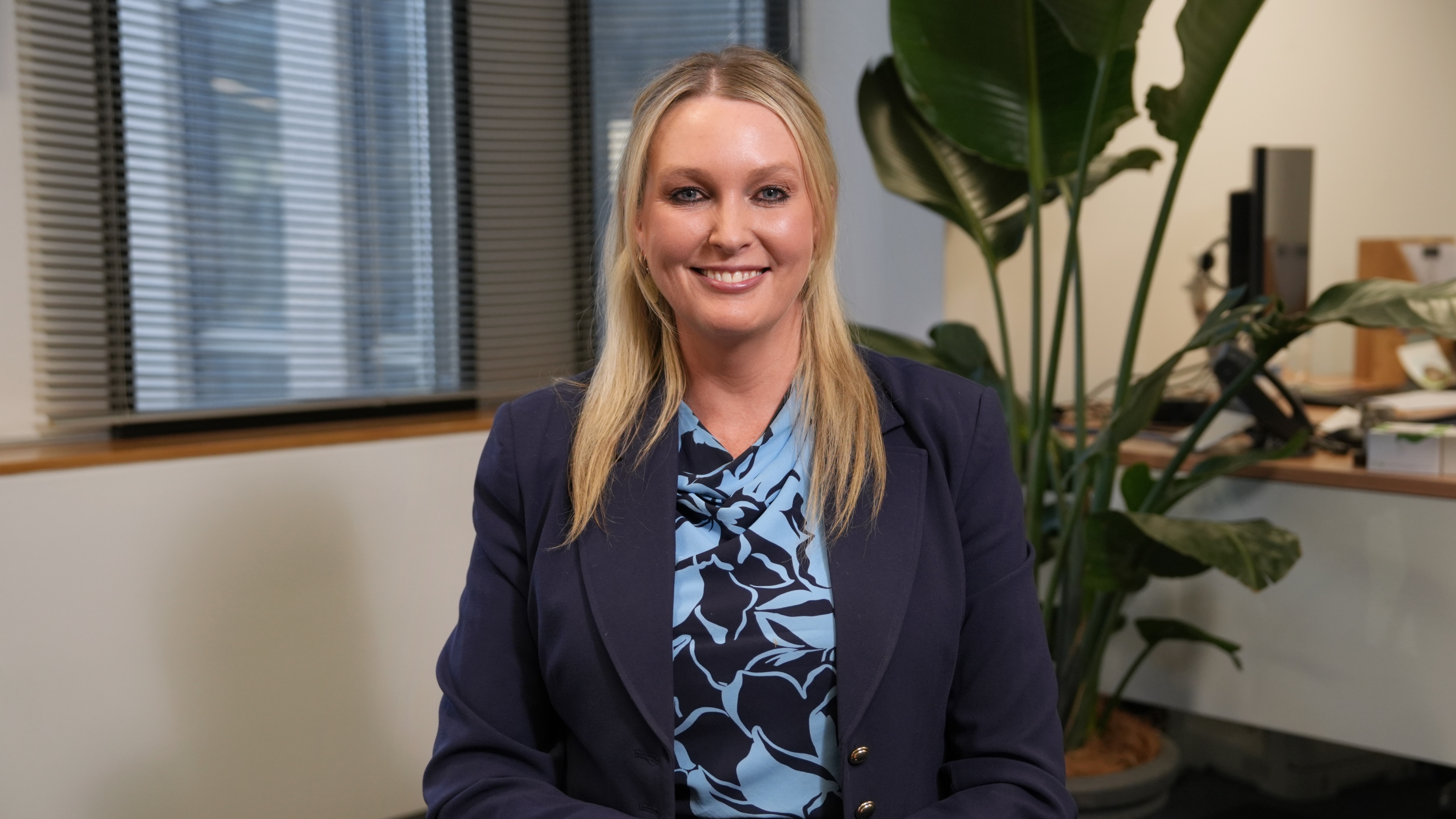 A woman with blonde straight hair in a blue blazer and top sits in an office smiling.