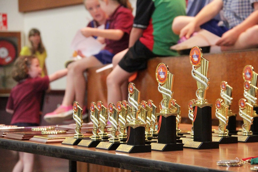 Trophies on a table in the foreground with children on a stage in the background.