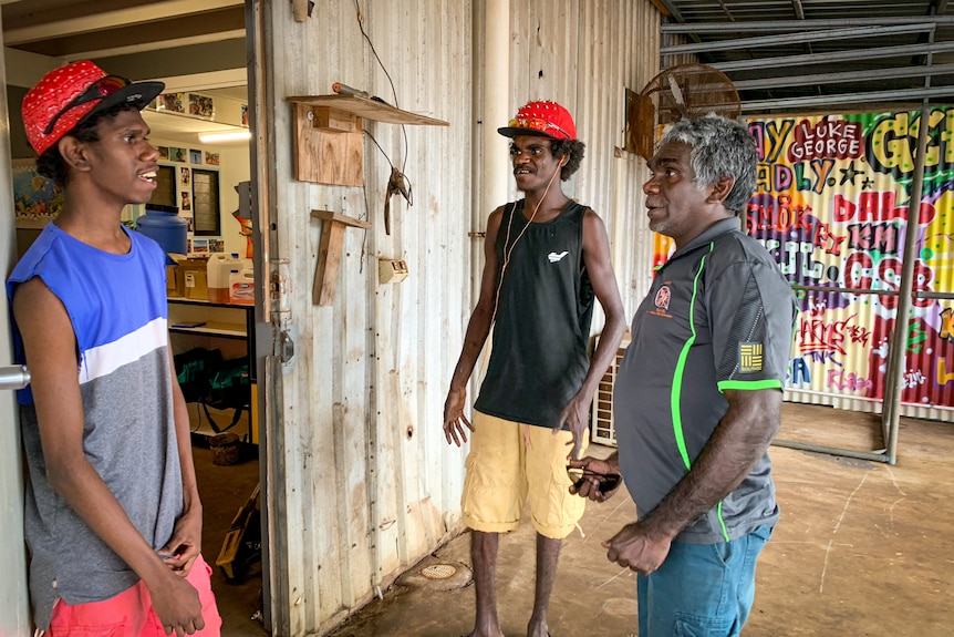 Young man Donovan Wurramara, left wearing a baseball cap, and Fabian Lalara, an older mand, stand outside a shed talking.