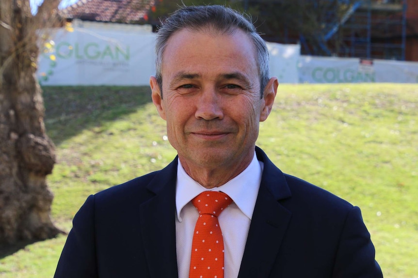 A head and shoulders shot of WA Health Minister Roger Cook smiling and standing outdoors wearing a suit and tie.