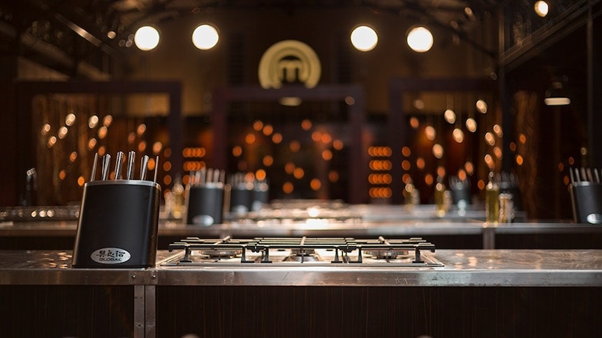 A dimly lit room showing rows of kitchen work benches featuring a gas stove and black knife block.