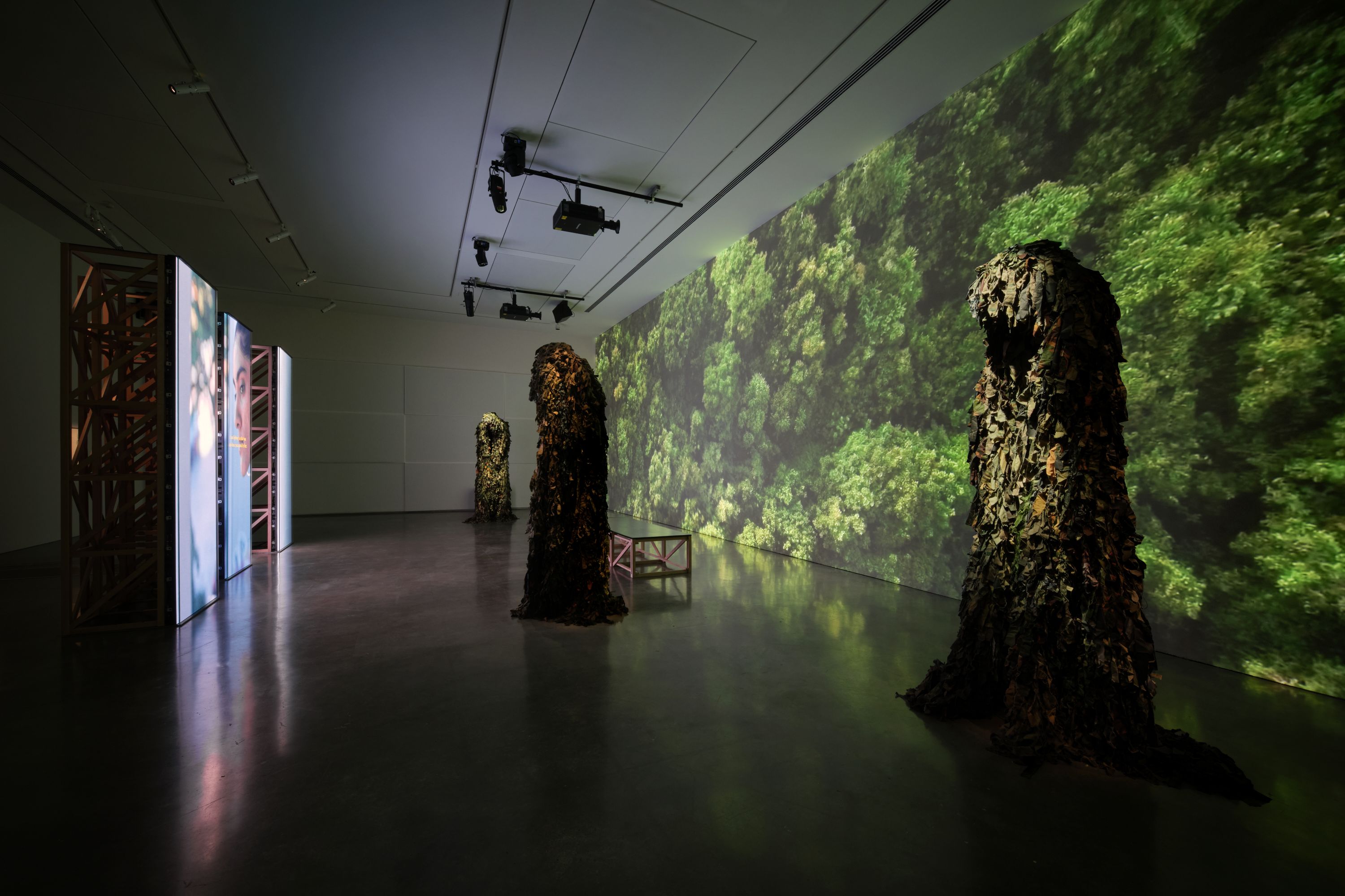 Duas grandes esculturas em frente a um grande telão mostrando uma vista aérea de uma copa verdejante de floresta; telas menores enfrentam isso
