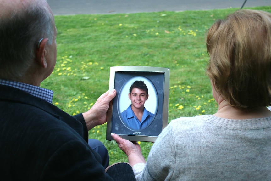 A man and a woman sit on a park bench holding a photo of a boy.