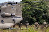 Soldiers exit a helicopter onto a field with long grass.
