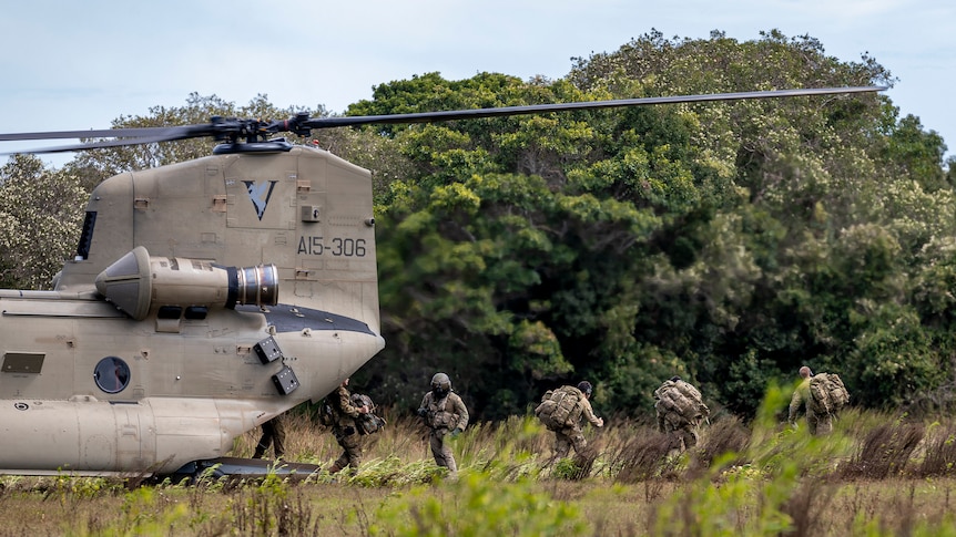 Soldiers exit a helicopter onto a field with long grass.