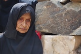 An old woman wearing black traditional dress leans against a rock wall.