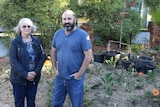 An elderly woman and a bearded man smile in front of a garden
