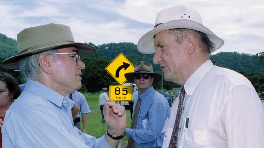 Two men wearing hats speak on the side of a rural highway, surrounded by media and minders