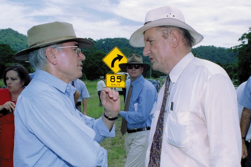 Two men wearing hats speak on the side of a rural highway, surrounded by media and minders