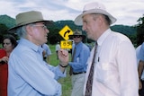 Two men wearing hats speak on the side of a rural highway, surrounded by media and minders