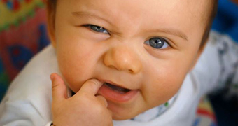 A baby looks up with one squinted eye while chewing on one finger.