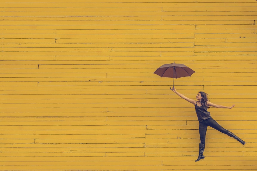A woman leaps with an umbrella in hand against a yellow wall.