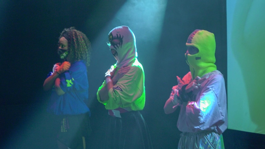 Three women on stage in ski masks with their arms crossed