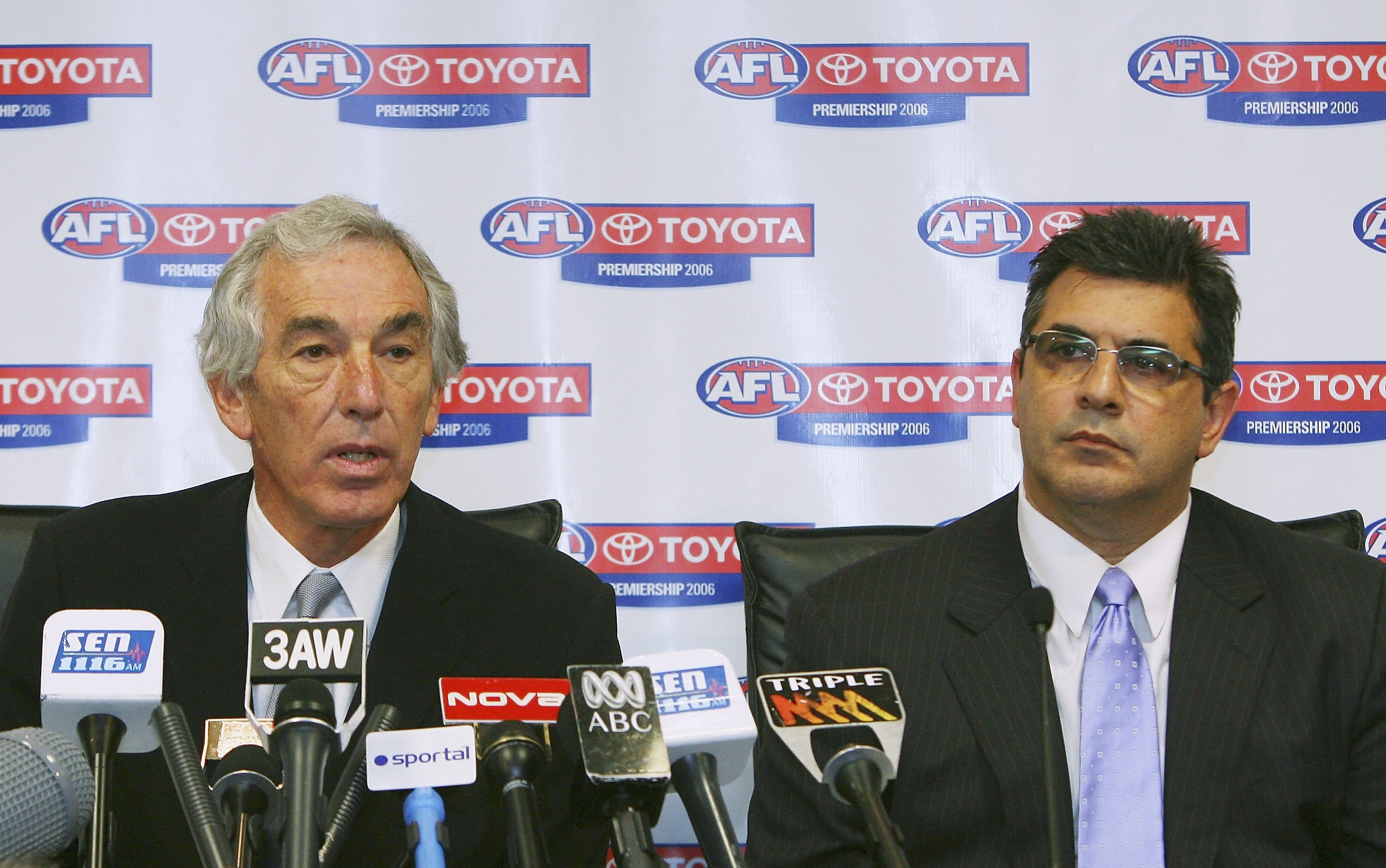 Two senior AFL executives sit in front of a bank of microphones, explaining an AFL Commission decision to the media.