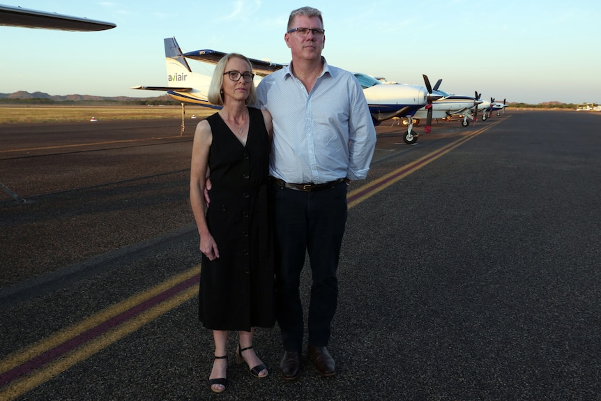 A man and woman stand on an airport taxiway in front of several light aircraft.
