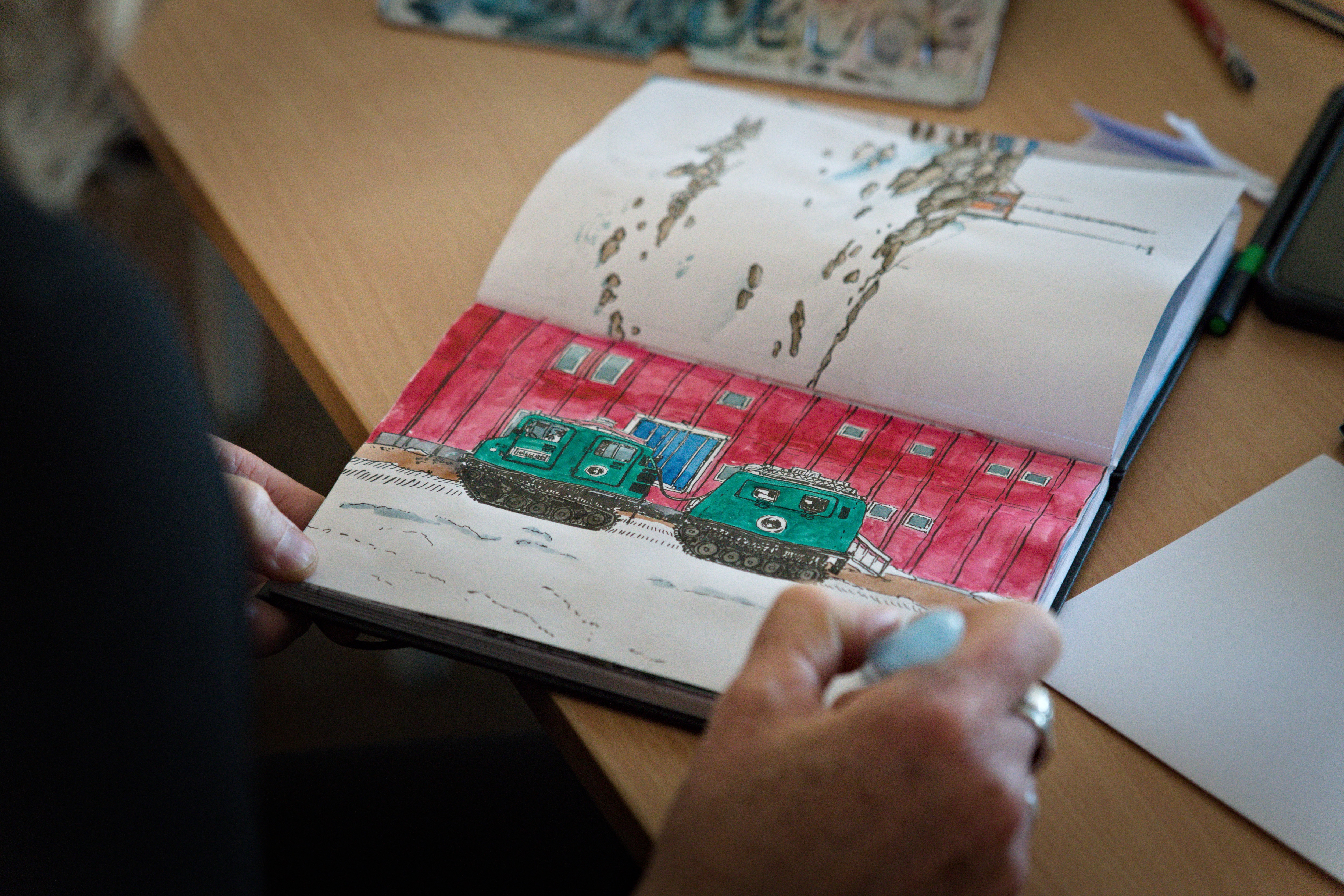 Um desenho a caneta e tinta de um edifício vermelho e dois veículos verdes em um caderno de desenho.