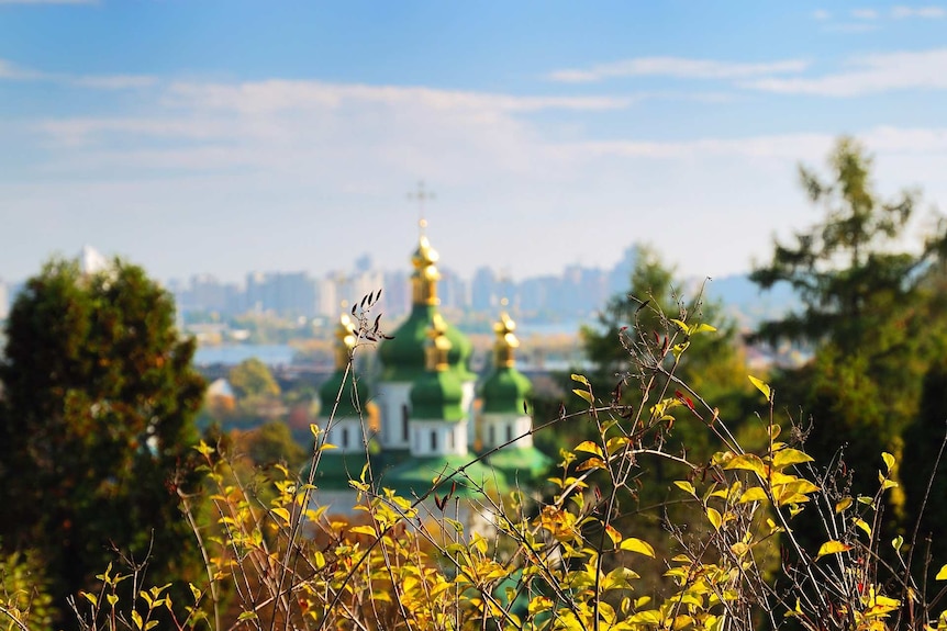 Foreground of grass and trees with blurred view of a green and gold monastery, and then a distant view of a city skyline.