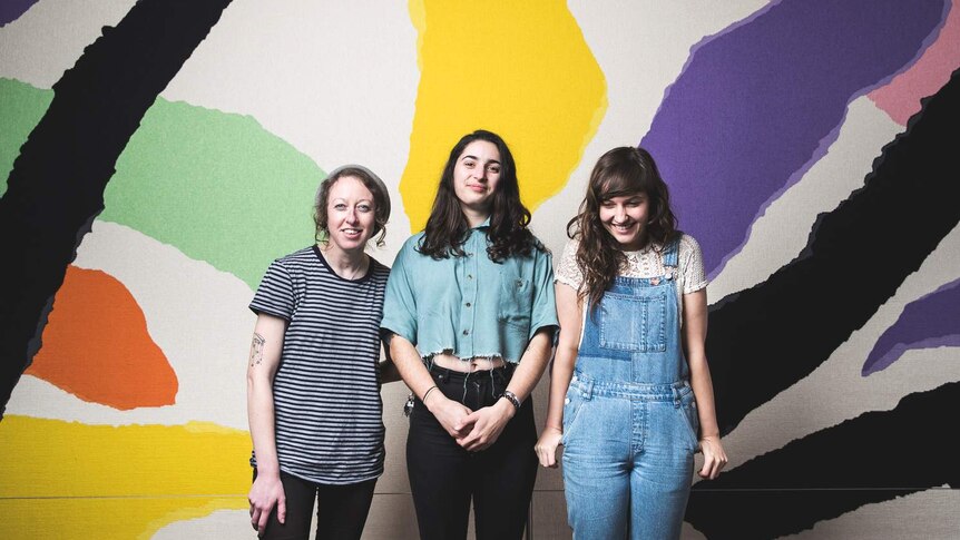 Camp Cope stand smiling in front of a splashed colour mural.