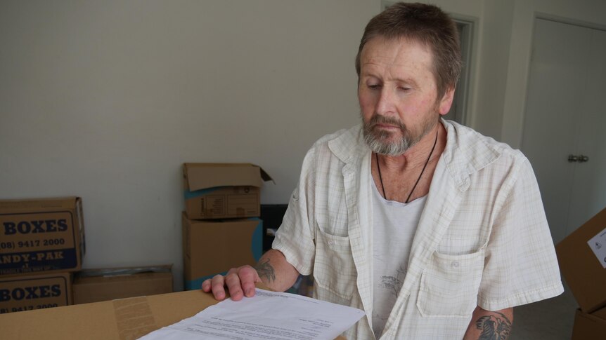 A sad-looking older man sits at a table, looks at a document, cardboard boxes behind him.