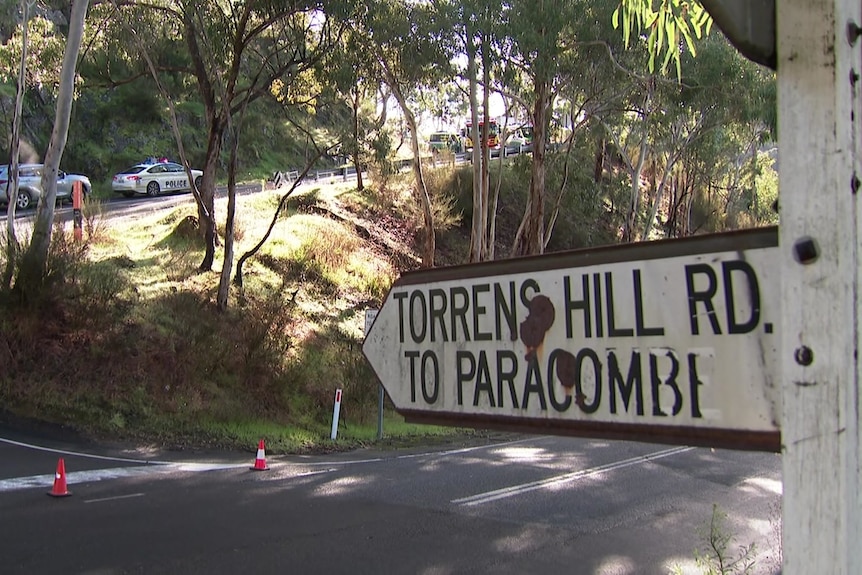 A sign reading 'Torrens Hill Rd to Paracombe'. In the background are traffic cones and emergency services vehicles