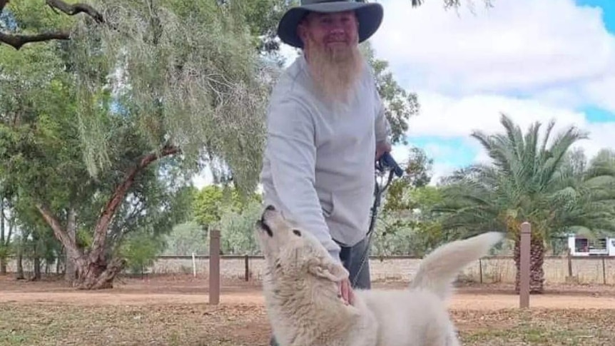 A man in a akubra style hat smiles at the camera while patting his white husky dog