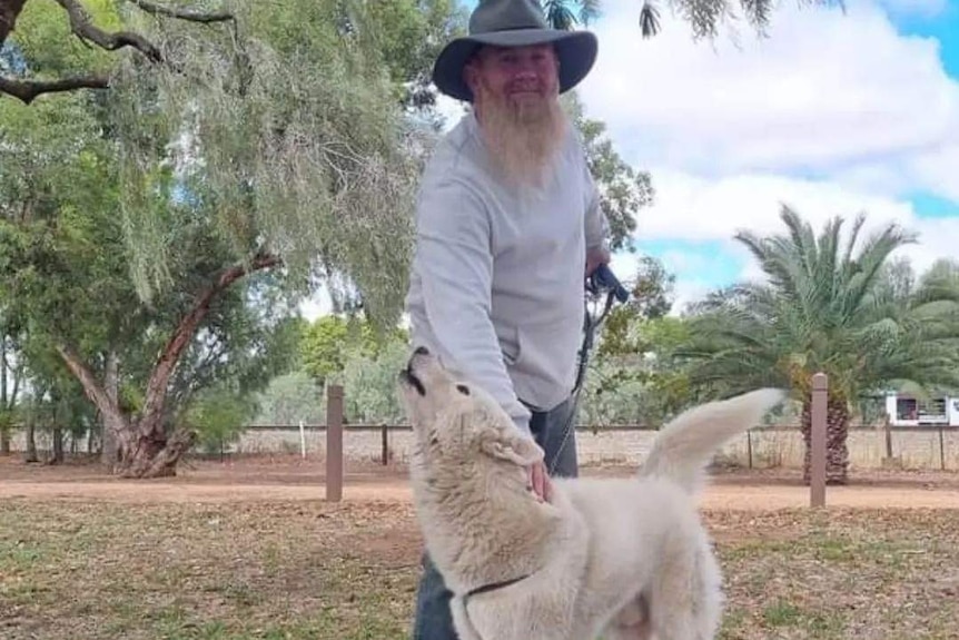 A man in a akubra style hat smiles at the camera while patting his white husky dog