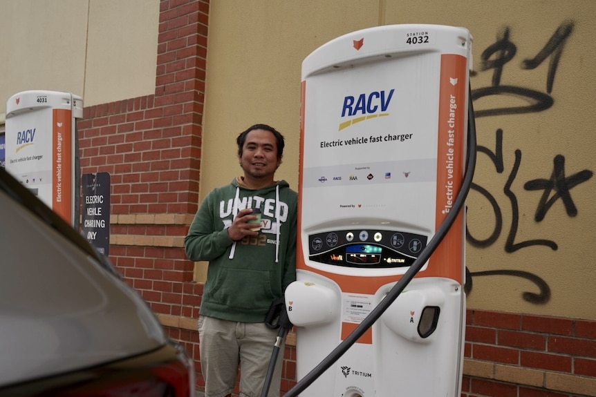 A man leans against an EV charger while holding a cup of coffee.