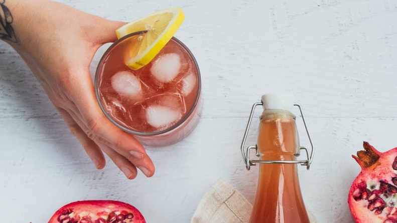 A woman holds a drink with ice on a table, next to a bottle for a story about kombucha and other "health" drinks.