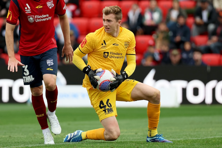 A blonde man wearing a yellow top and shorts kneels down on grass and holds a white soccer ball