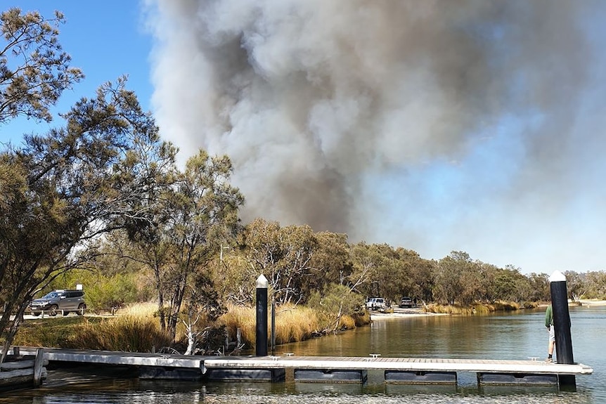 thick smoke billows into the sky from a bushfire with trees, a lake and jetty below.