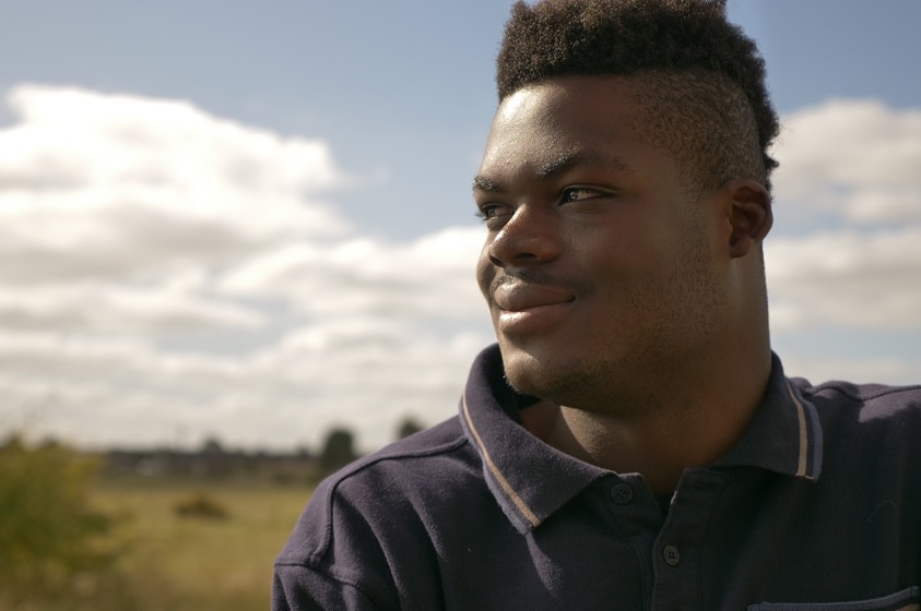 A young man, Lunor Folly, looks to the left of the frame, with a blue sky and green landscape behind him.