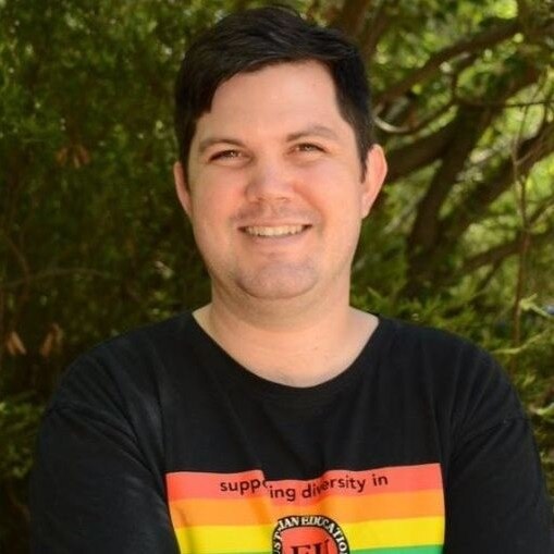 A man in a T-shirt with a rainbow logo celebrating diversity, smiles.