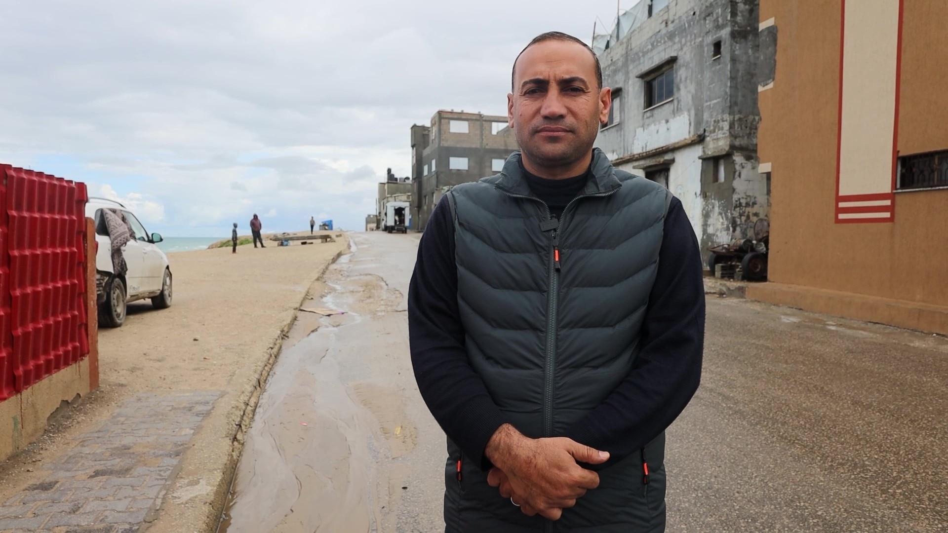 a man in a puffer vest jacket standing on the road near a beach and damaged buildings