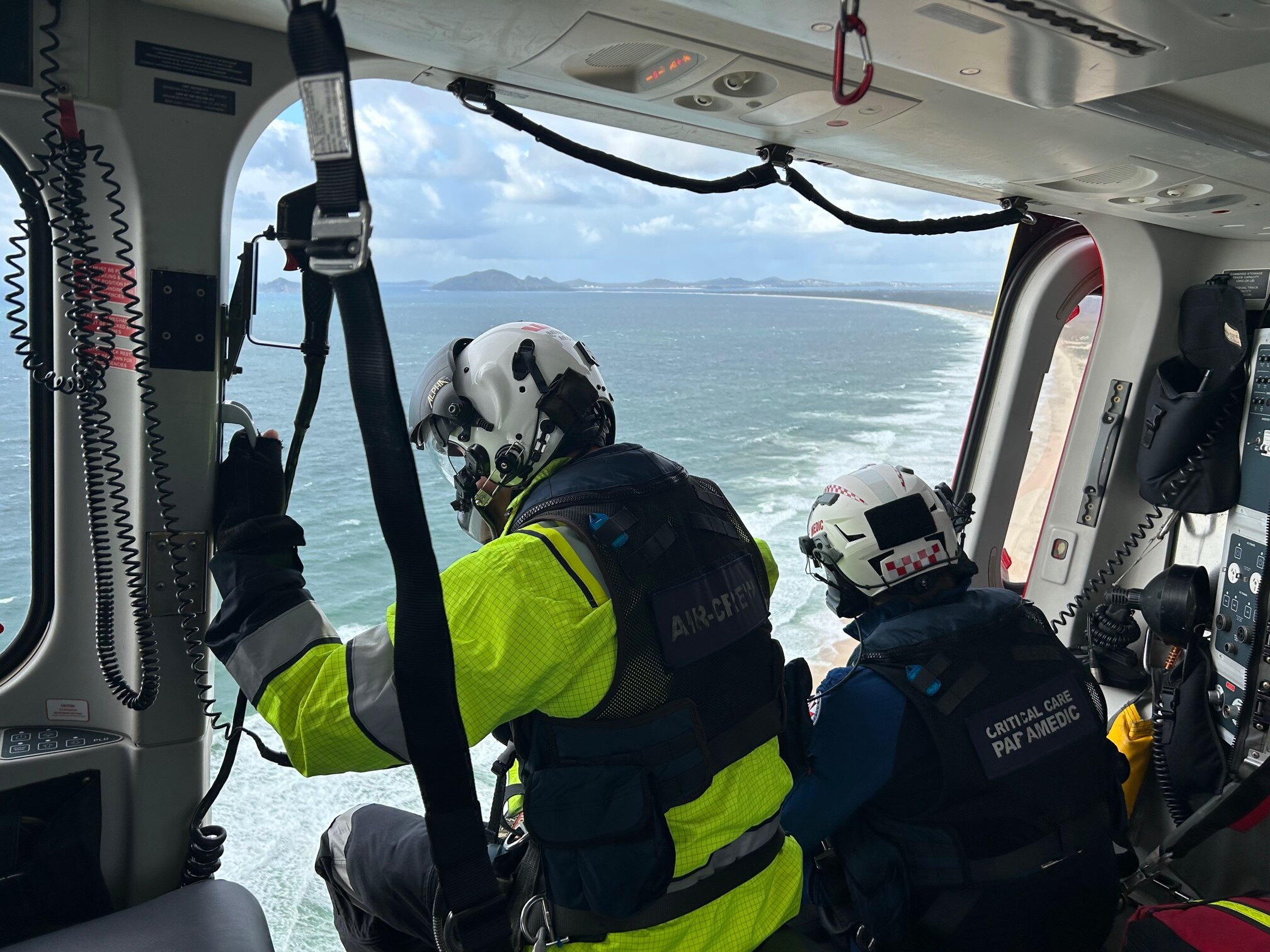 Two people in high vis in a helicopter flying over water 