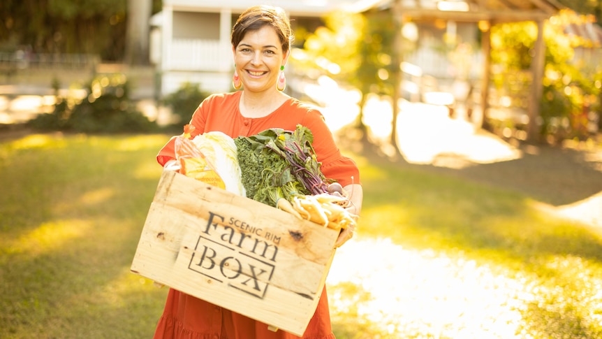 Smiling short-haired woman in orange dress, holds a produce crate with Farm Box stenciled on it, stands in sunny grassy area.