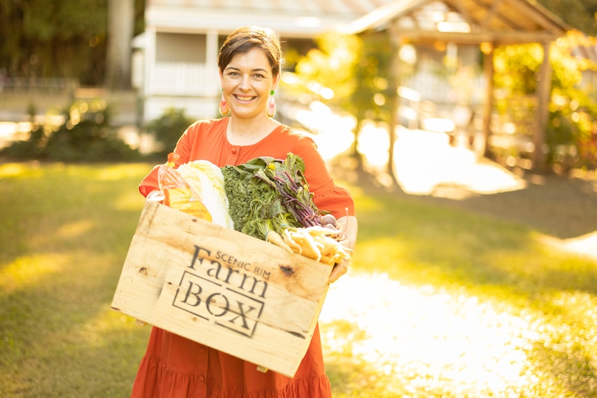 Smiling short-haired woman in orange dress, holds a produce crate with Farm Box stenciled on it, stands in sunny grassy area.
