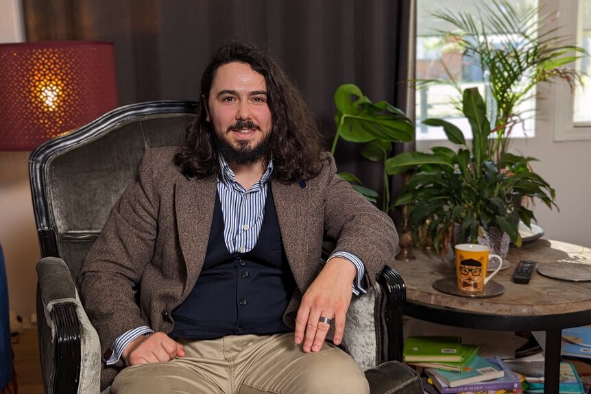 A man with shoulder-length dark curly hair and facial hair sits in an armchair in a maximalist apartment.