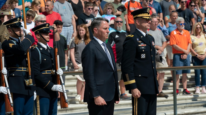 A white man in a suit stands in front of a crowd with several military officers in Arlington National Cemetary
