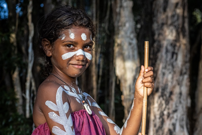 Girl wearing violet dress and white paint holding stick in a forest.