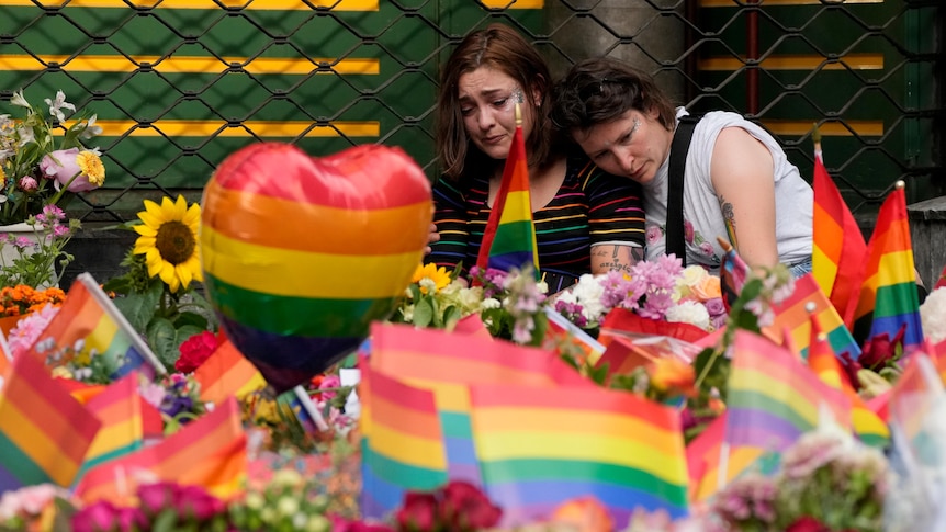 People comfort each other at the scene alongside flowers and rainbow flags