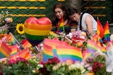 People comfort each other at the scene alongside flowers and rainbow flags