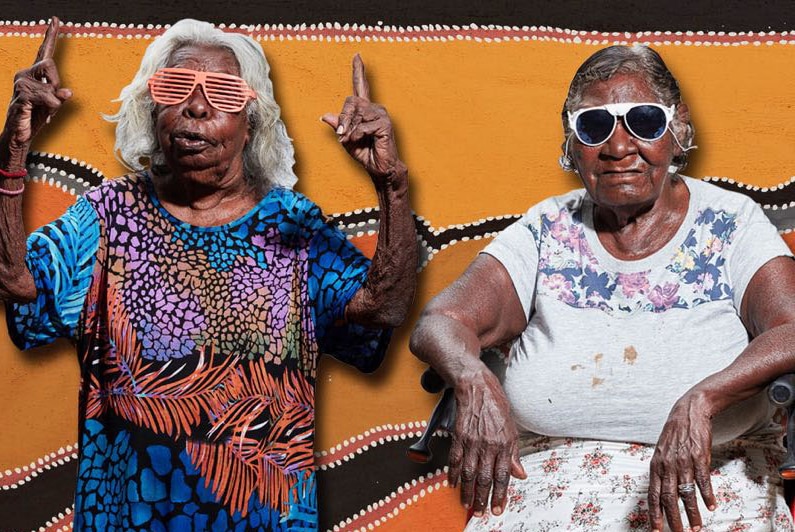 Two Aboriginal women wearing sunglasses, one seated and one with raised hands,  in front of a dot painting.